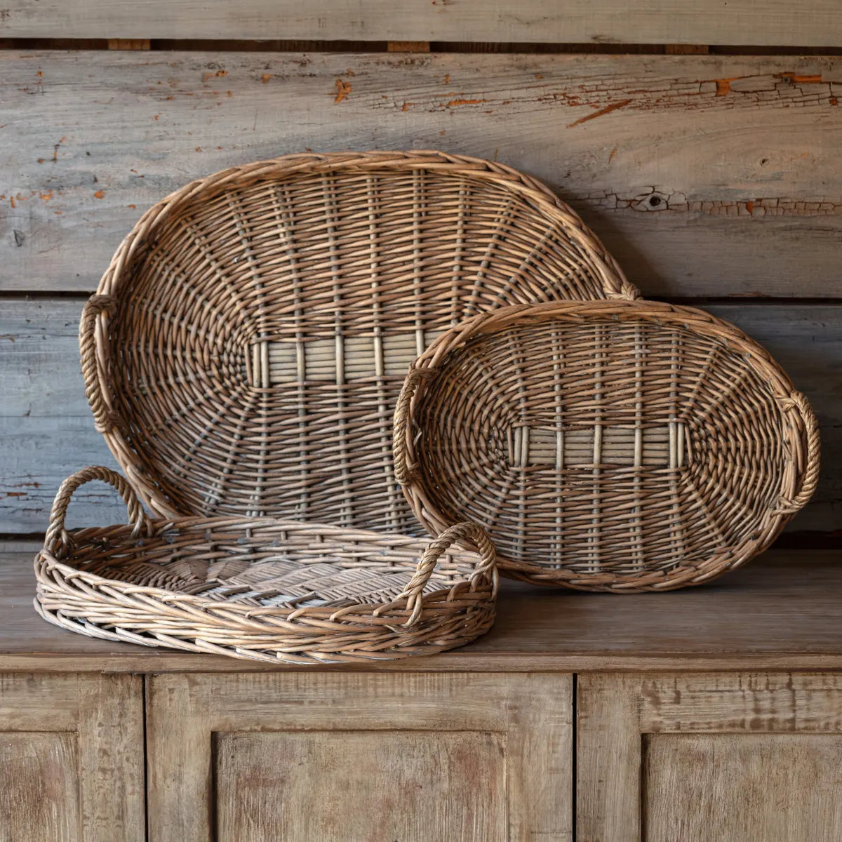 Three oval hand woven wicker basket trays with handles against a wooden surface with a rustic wooden wall background. 