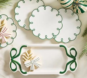 White ceramic holiday party serving pieces with green scalloped borders, including a rectangular platter and round plates, styled with decorated cookies and pine branches on a light marble surface.