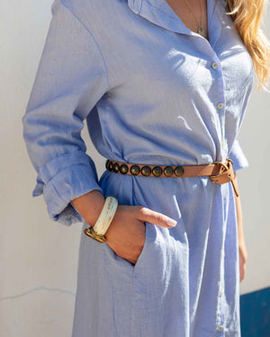 model wearing a light blue linen shirt dress with a brown belt against a neutral background. 