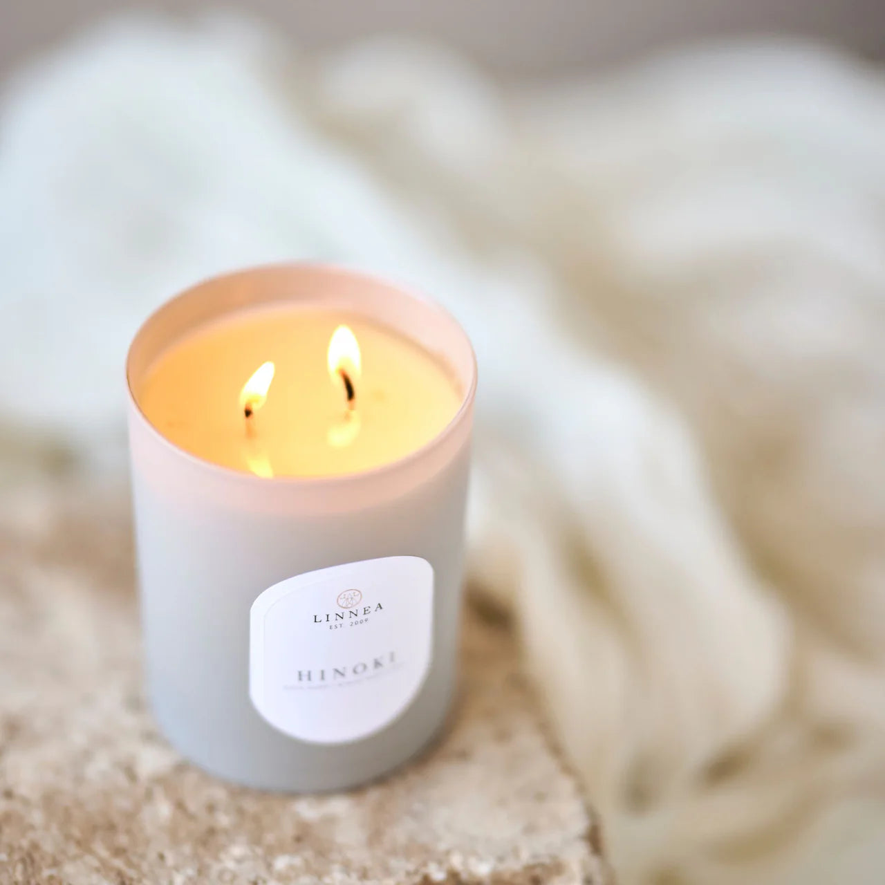 Lit scented candle in "Hinoki" with visible brand label "Linnea"on a blurred textured background. 