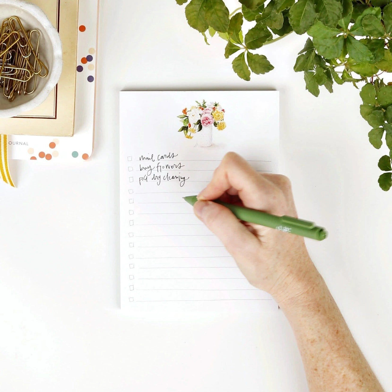 Hand writing on a checklist notepad with a pen, surrounded by stationery items and a plant. Emily Lex Studios. 