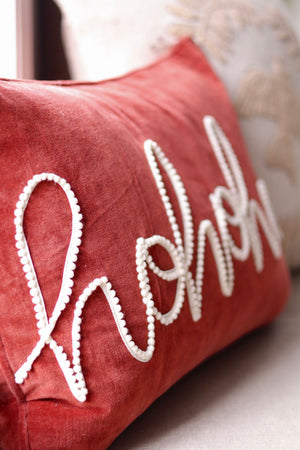 A dusty wine red velvet throw pillow featuring the word 'ho ho ho' stitched in white cursive with beaded outlines, resting on a light-colored sofa.