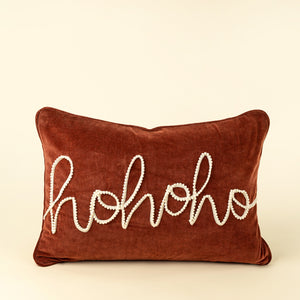 A rust red velvet holiday lumbar pillow featuring the word 'ho ho ho' embroidered in white cursive across the front, set against a solid cream background.