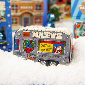 A festive gray box of MAeve chocolate bonbons shaped like a Christmas camper, sitting in white faux snow with blurred colorful holiday decorations in the background.