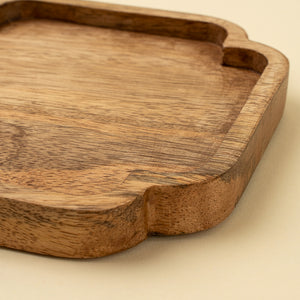 A close-up of a scalloped wooden serving tray with a warm medium-brown finish and visible wood grain, set against a light beige background.