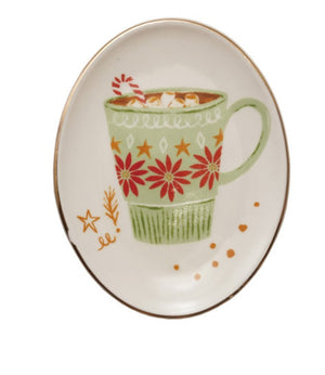 An oval white ceramic plate with a gold rim, featuring a hand-painted illustration of a green mug filled with hot cocoa, marshmallows, and a candy cane, decorated with red flowers and gold stars.