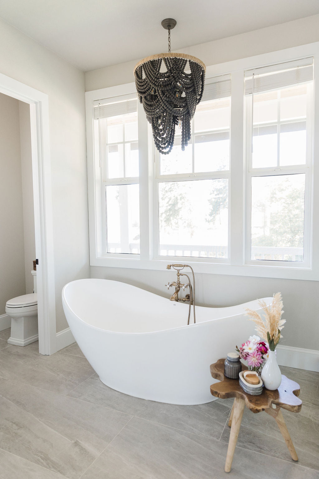 White stand alone tub with wood side table and floral arrangement and a dramatic gray beaded chandelier