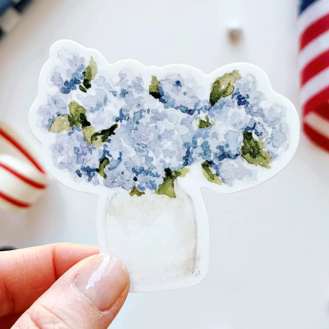 Hand holding a sticker with a blue floral hydrangea design on a blurred background. Emily Lex. 