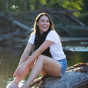 A woman laughing while sitting on a large log by a body of water, wearing a white t-shirt, a black scarf, and denim shorts.