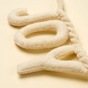 Cream-colored felt letter banner spelling 'JOY' with a braided cord, shown on a matching cream background.