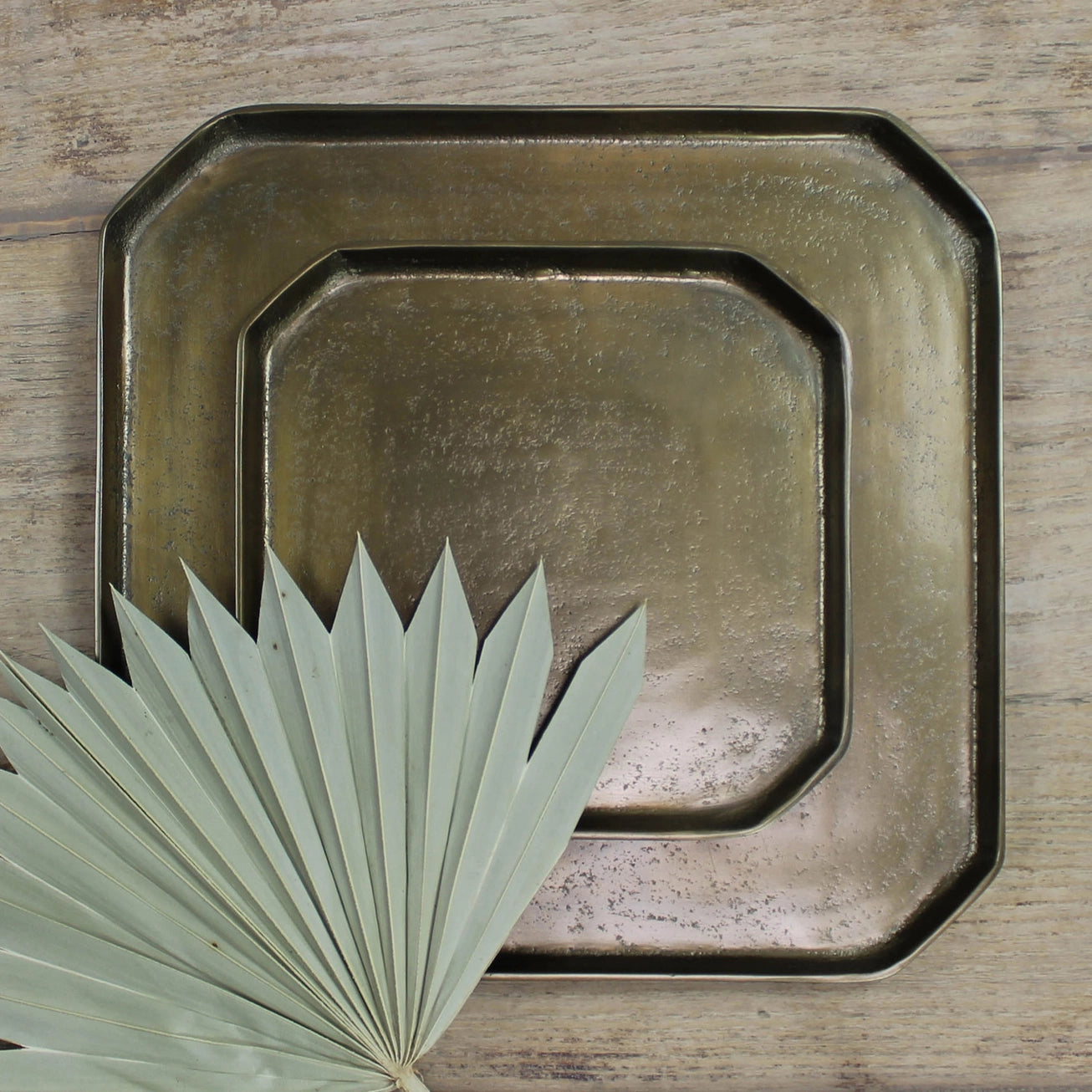 Set of two large and small square brass trays on a wooden surface with a palm leaf. 