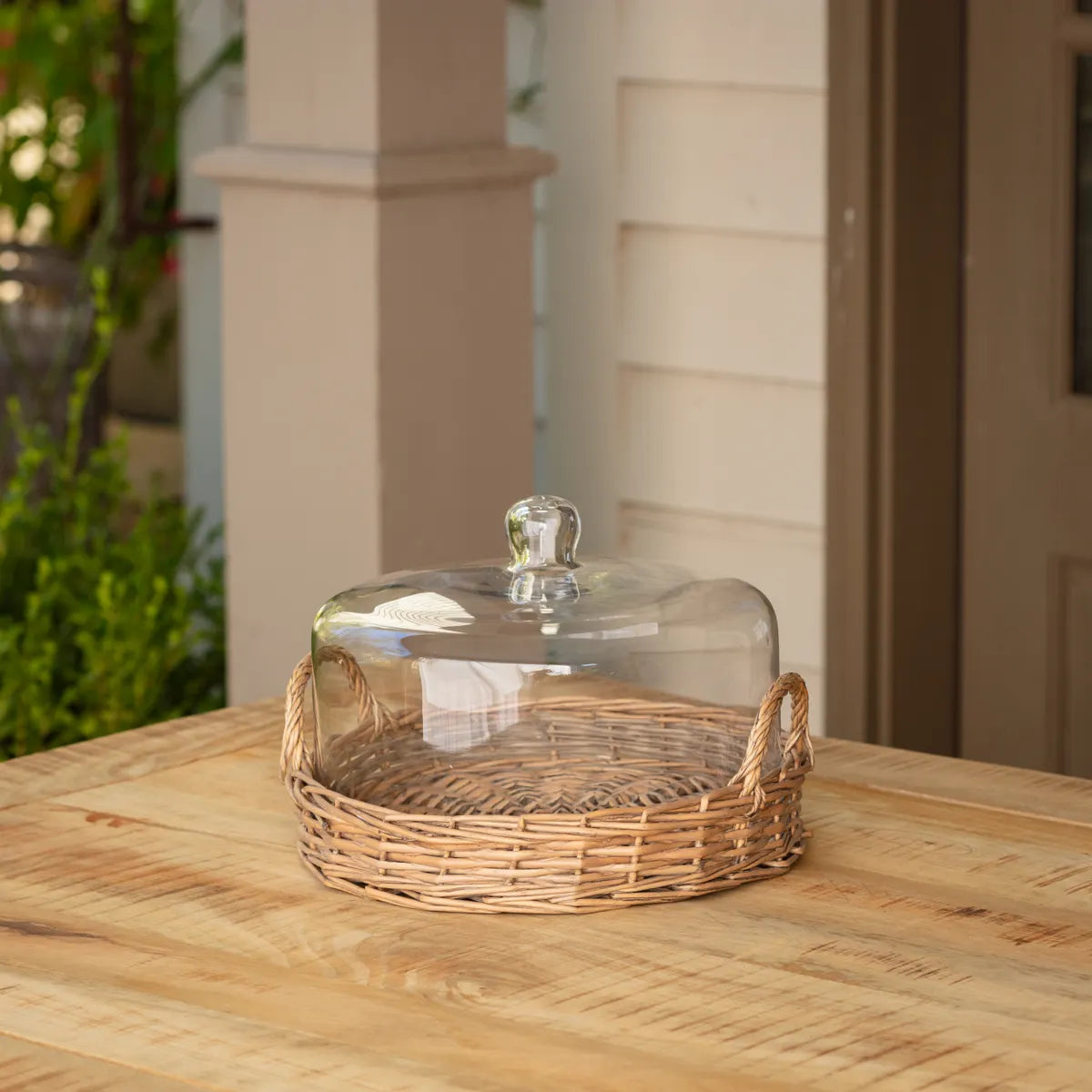Wicker basket with glass cloche lid on a wooden outdoor table surface. Farmhouse, coastal, and french inspired kitchen serveware. 