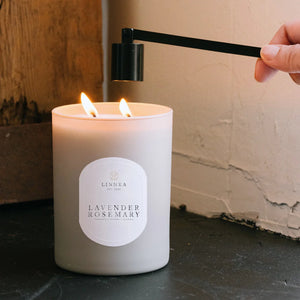Hand holding candle snuffer while lit, featuring 'Lavender Rosemary' label on a neutral background.