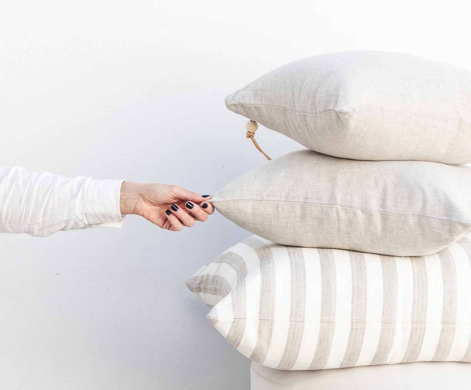 Collection of oatmeal colored linen and down pillows stacked against wall. 