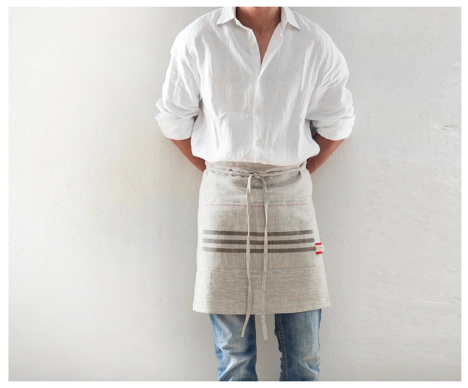 Male model wearing half waist linen kitchen apron with horizontal blue stripe. Tied around midline. Linen apron with pockets.