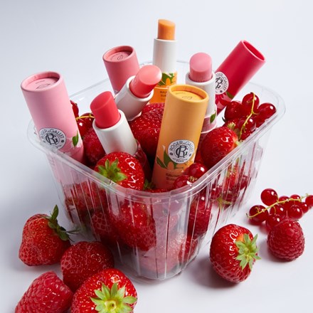 A clear plastic tub filled with assorted lip balm tubes in pink, coral, peach, and red tones, surrounded by fresh strawberries and red berries on a white background.