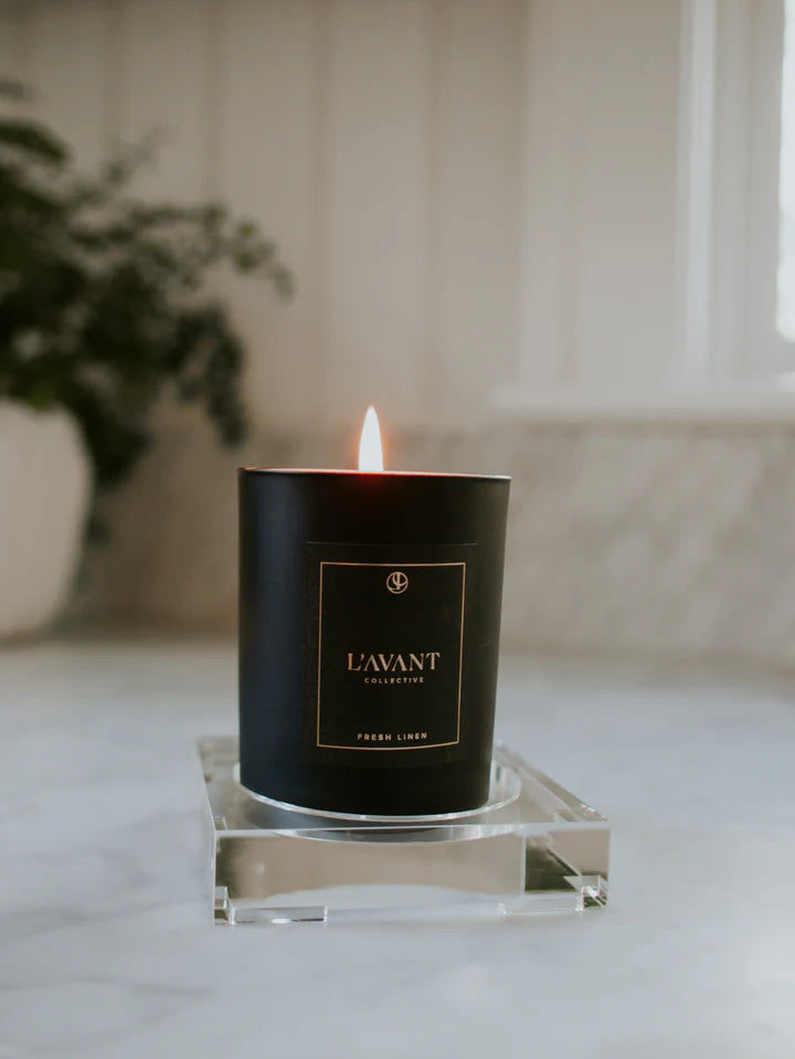 Matte black fresh linen candle labeled L"AVANT on clear acrylic tray with blurred background. 