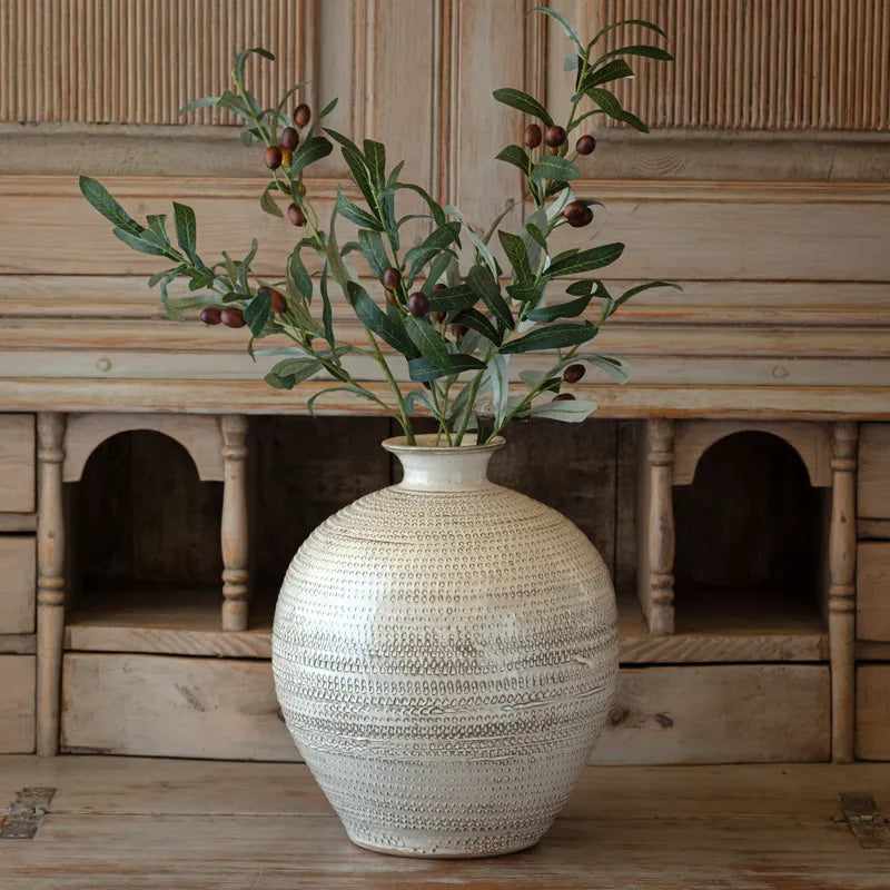 Textured terracotta urn style vase with olive stem greenery on a wooden surface. 