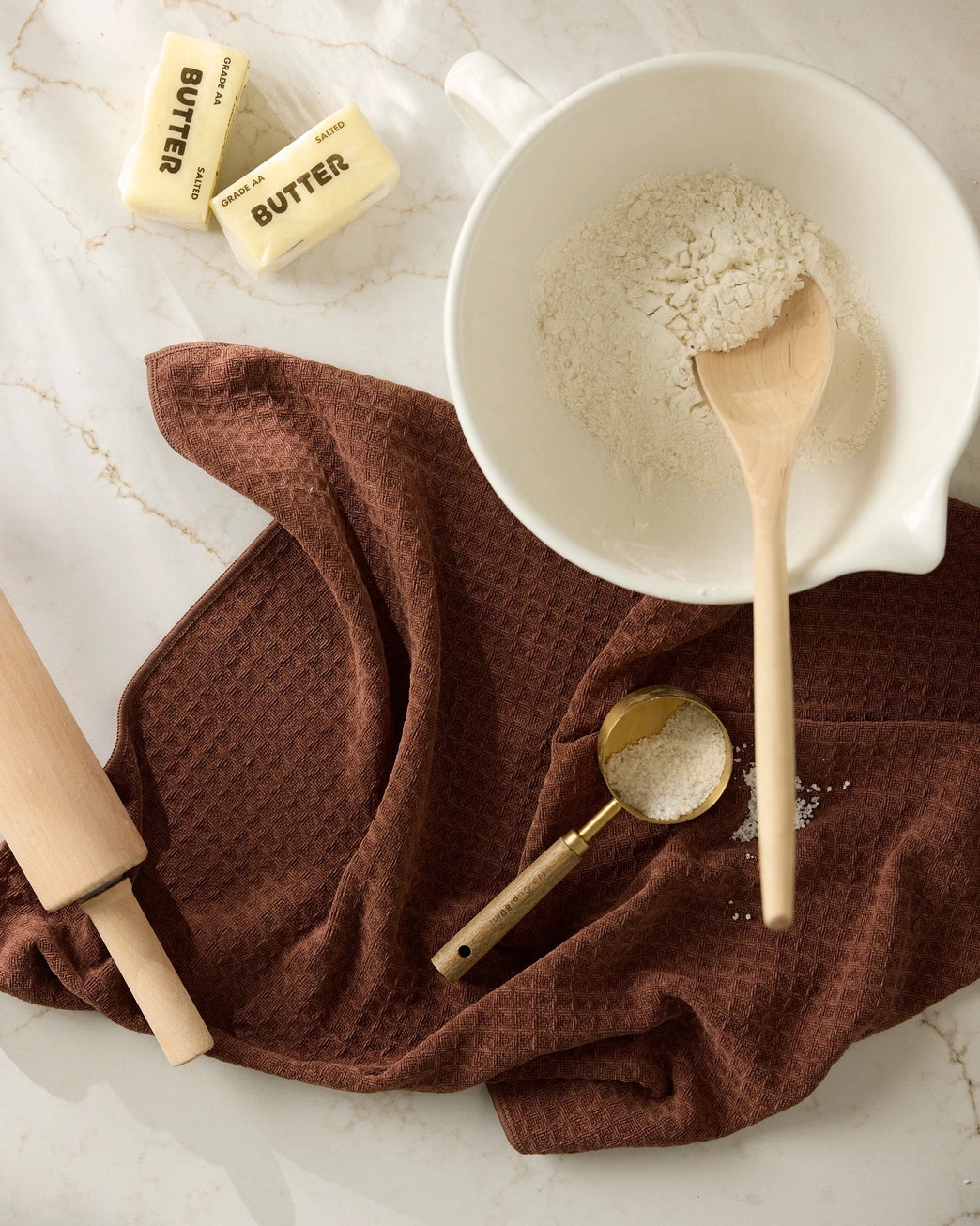Bowl of flour with a wooden spoon, rolling pin, and sticks of butter on a maple brown waffle weave microfiber hand towel. Geometry House. 