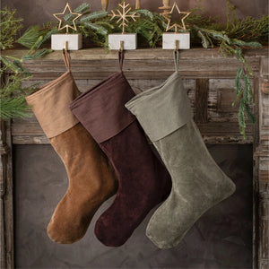 Three Christmas stockings in brown, burgundy, and sage green suede hang from a rustic wooden mantel decorated with pine branches and gold star ornaments.