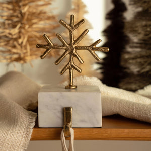 A gold snowflake-shaped hanging ornament mounted on a white marble cube base with a small hook and cord, styled on a wooden surface with soft neutral textiles and blurred holiday decor in the background.