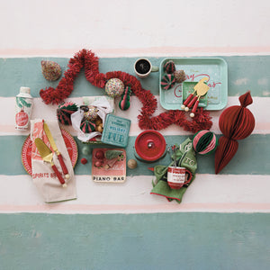 A styled flat-lay of vintage Christmas-themed items arranged on a white and mint green striped surface. The collection includes red tinsel garland, red and green striped ornaments, a mint green tray with a Santa figure, a red candle, a small 'Piano Bar' tin, a red-and-white patterned napkin, and other holiday accessories.