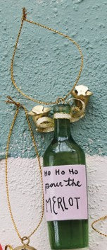 A green glass bottle pendant necklace with a gold-tone chain and decorative gold charms, featuring a pink handwritten label that reads 'HO HO HO pour the MERLOT'.