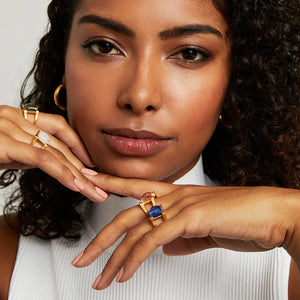 Woman wearing multiple mini plaza gold rings featuring different colored gemstones on a plain background. Dean Davidson jewelry. 