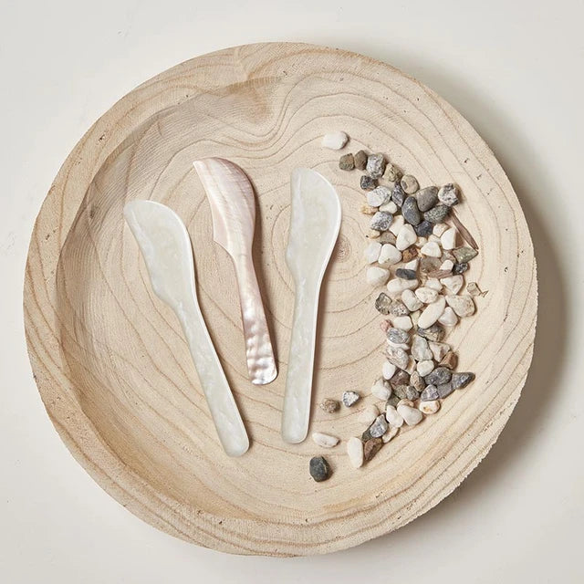 Three translucent mother-of-pearl cutlery pieces, including two spoons and a knife, arranged on a round light wood tray alongside small white and gray pebbles.