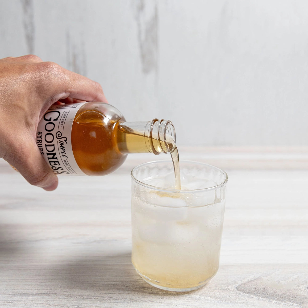 Hand pouring orange vanilla simple syrup from Simple Goodness Sisters into a glass on a light background.
