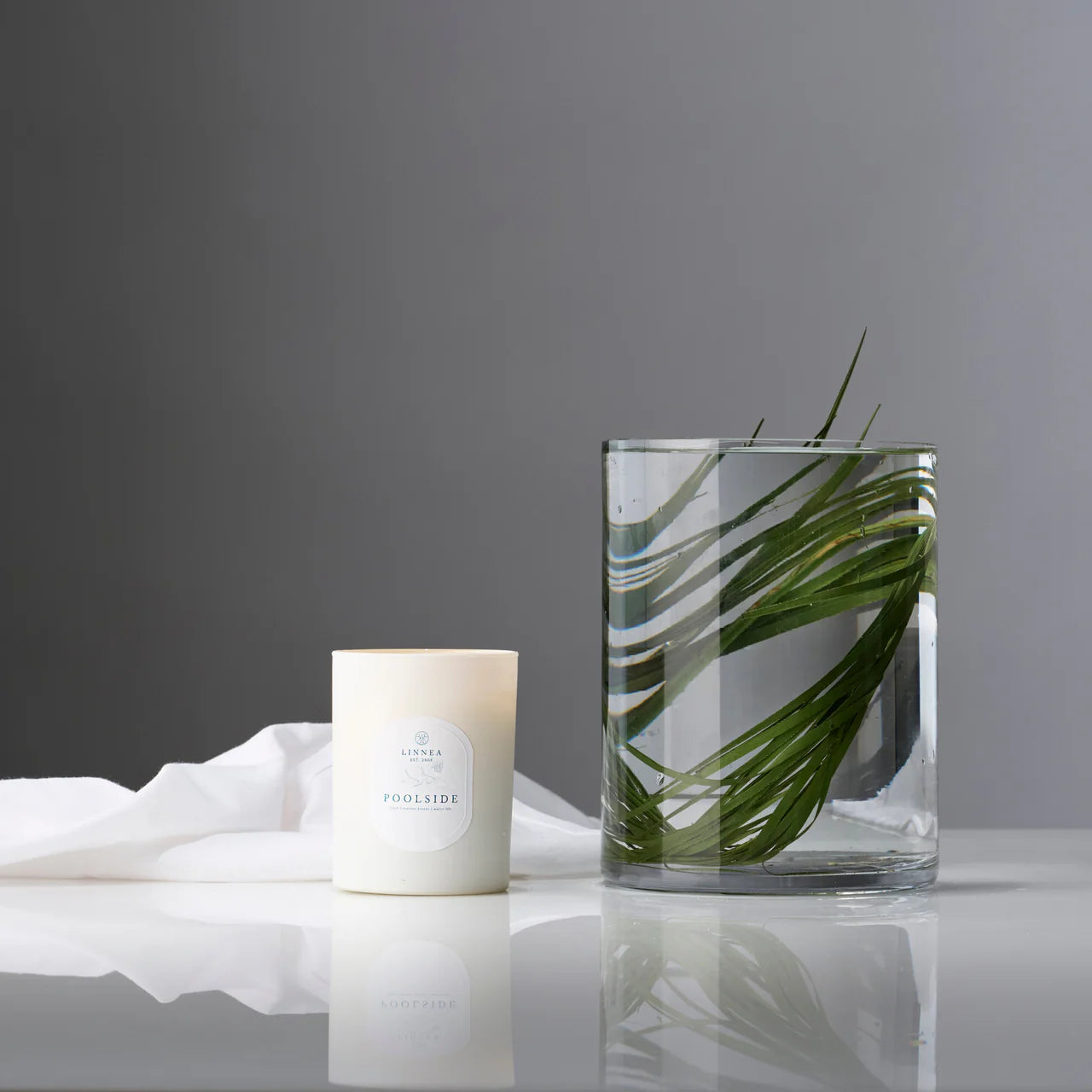 Poolside scented candle and glass votive filled with palm leaf on a reflective surface against a gray background. Linnea Co. 