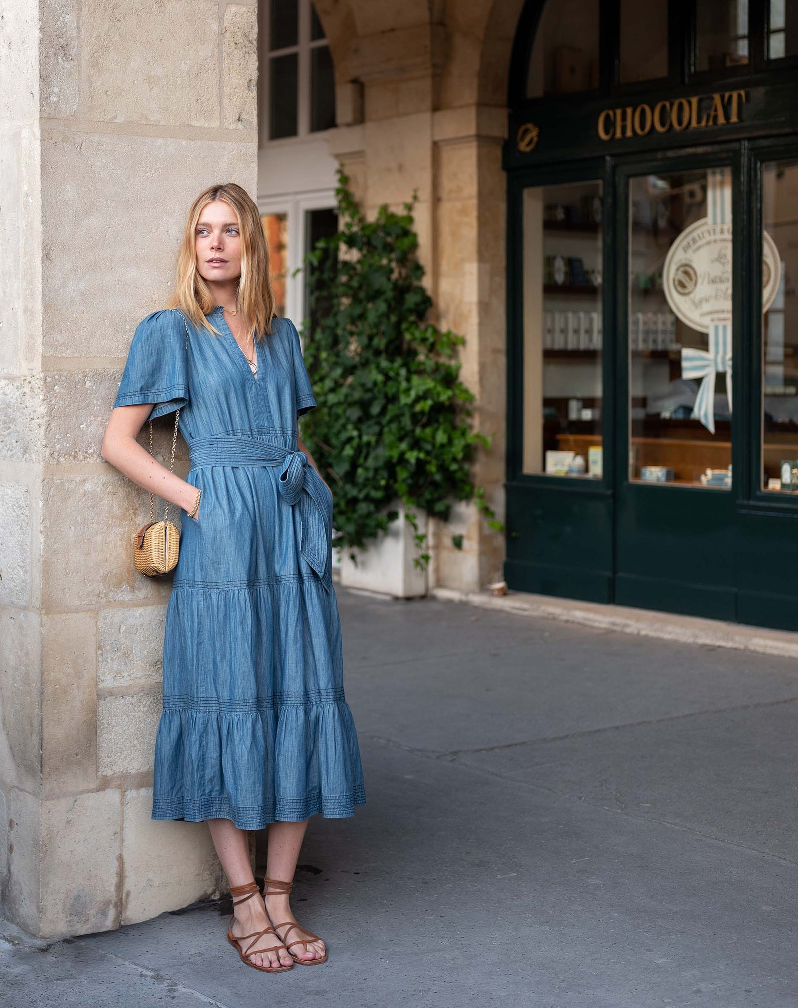 Woman in a blue maxi cotton dress with tiered ruffle design standing outside a building with 'Chocolat' sign.