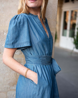 Woman wearing a blue dress with a stone wall and building in the background. Side profile view. 