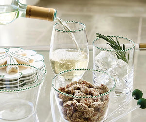 A set of clear glass partyware with green and white candy cane striped rims, including a wine glass being filled with white wine, a bowl of almonds, a bowl of olives, and a tumbler with ice and rosemary, on a glass table.
