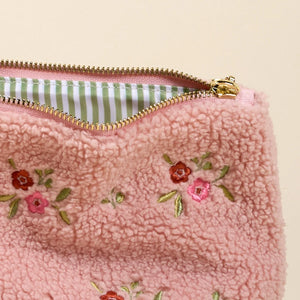 Close up detail  of light pink sherpa teddy cosmetics bag with embroidered red and pink flowers on the front. Bag is opened to display the striped green lining. 