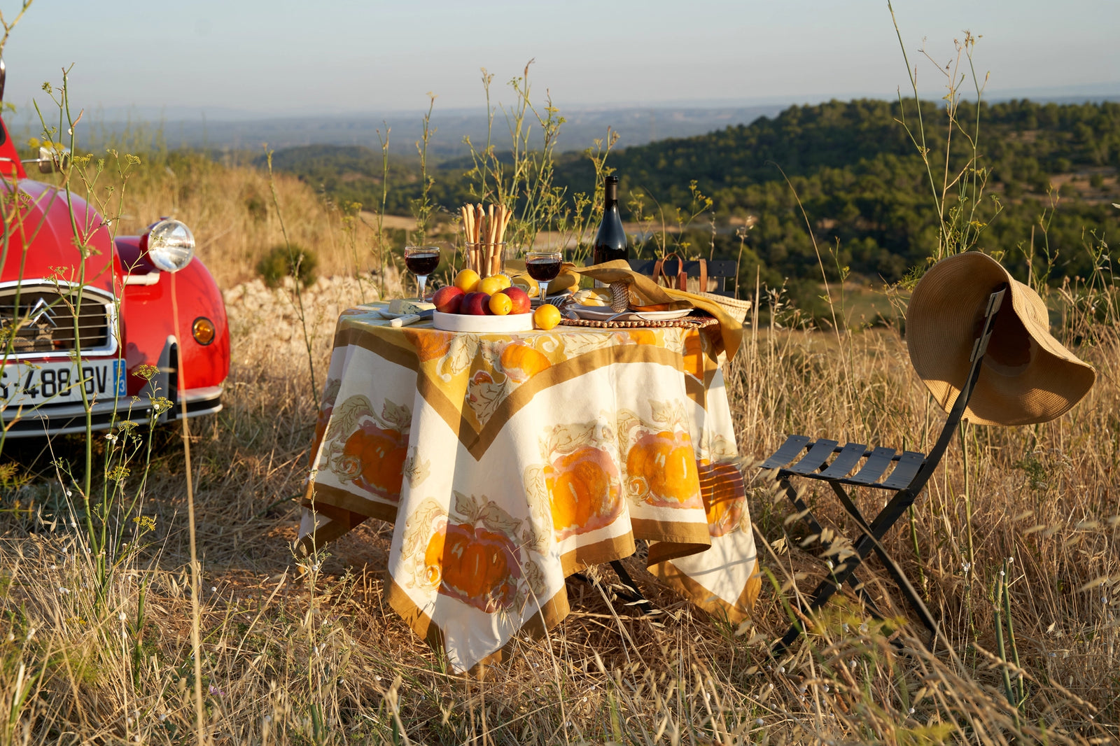 Autumn tablescape with seasonal fruits wine, and orange pumpkin cotton printed tablecloth; Set in a rustic countryside with an old red truck and trees in the distance. 
