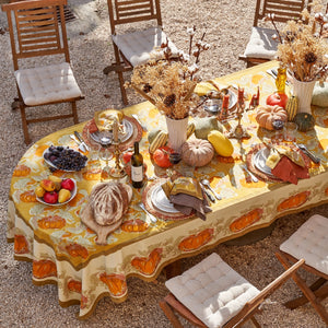 An outdoor autumn-themed table setting featuring a yellow tablecloth with a pumpkin and floral pattern, surrounded by wooden chairs with cream cushions. The table is decorated with fall produce, plates, wine, and dried cotton bolls in vases.