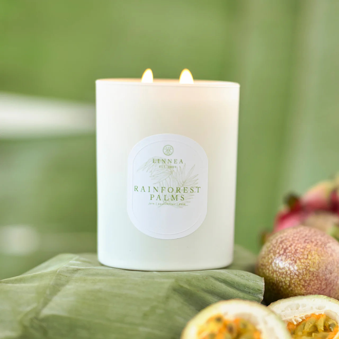 White candle labeled 'Rainforest Palms' on a green surface with blurred background and passion fruits  displayed below. 