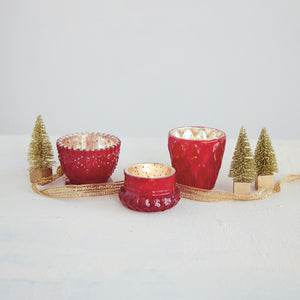 Three red textured glass candle holders with gold glitter interiors, arranged on a white surface with decorative gold ribbon and small gold tree ornaments against a light gray background.