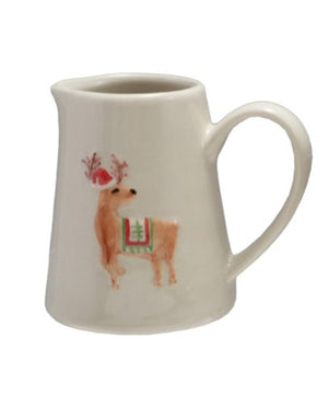 An off white ceramic creamer pitcher with a handle, decorated with a brown reindeer wearing a red Santa hat and a green Christmas sweater, on a plain white background.