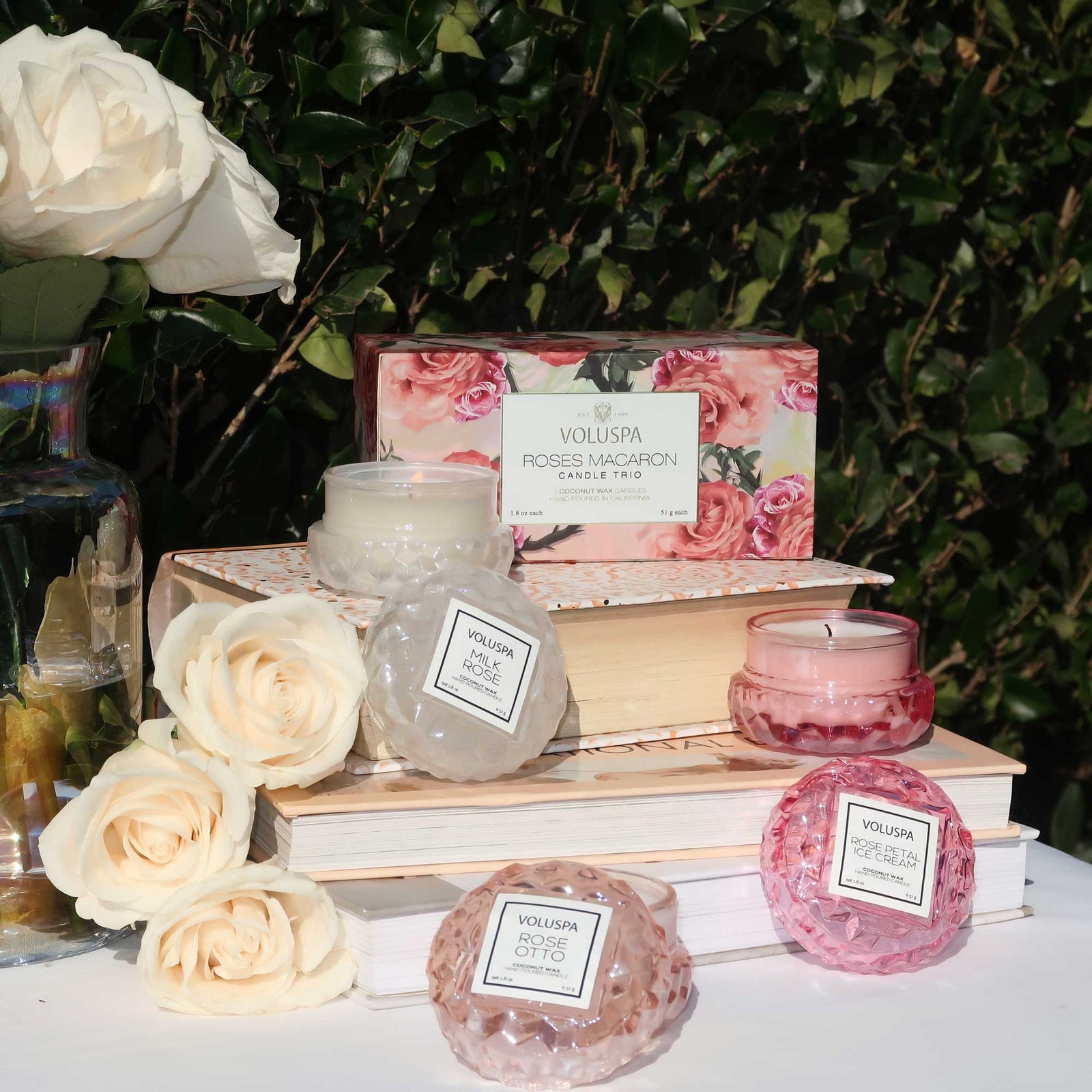 Decorative roses and pink colored books scattered on table with three macaroon glass candles displayed. Voluspa Rose Milk, Rose Otto, and Rose Petal Ice Cream. 