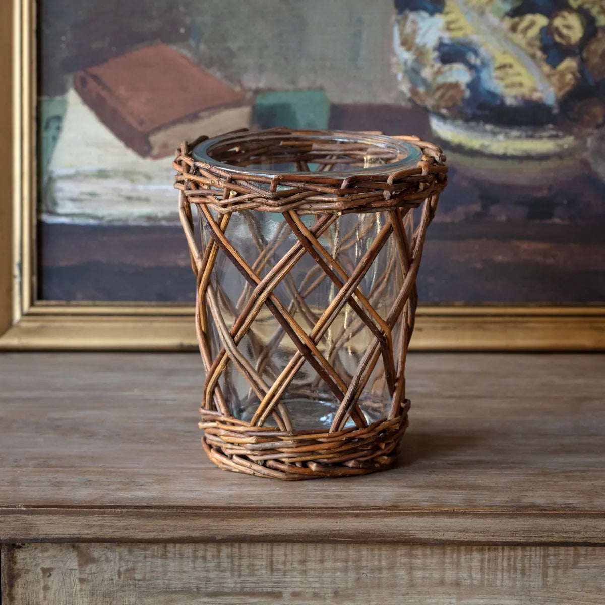 Wicker candle holder on a wooden surface with a painting in the background. 