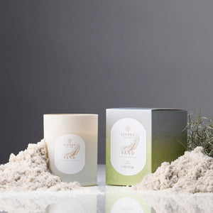 Sand scented candle on a textured surface with sand displayed against a dark background. Linnea soy wax candle. 