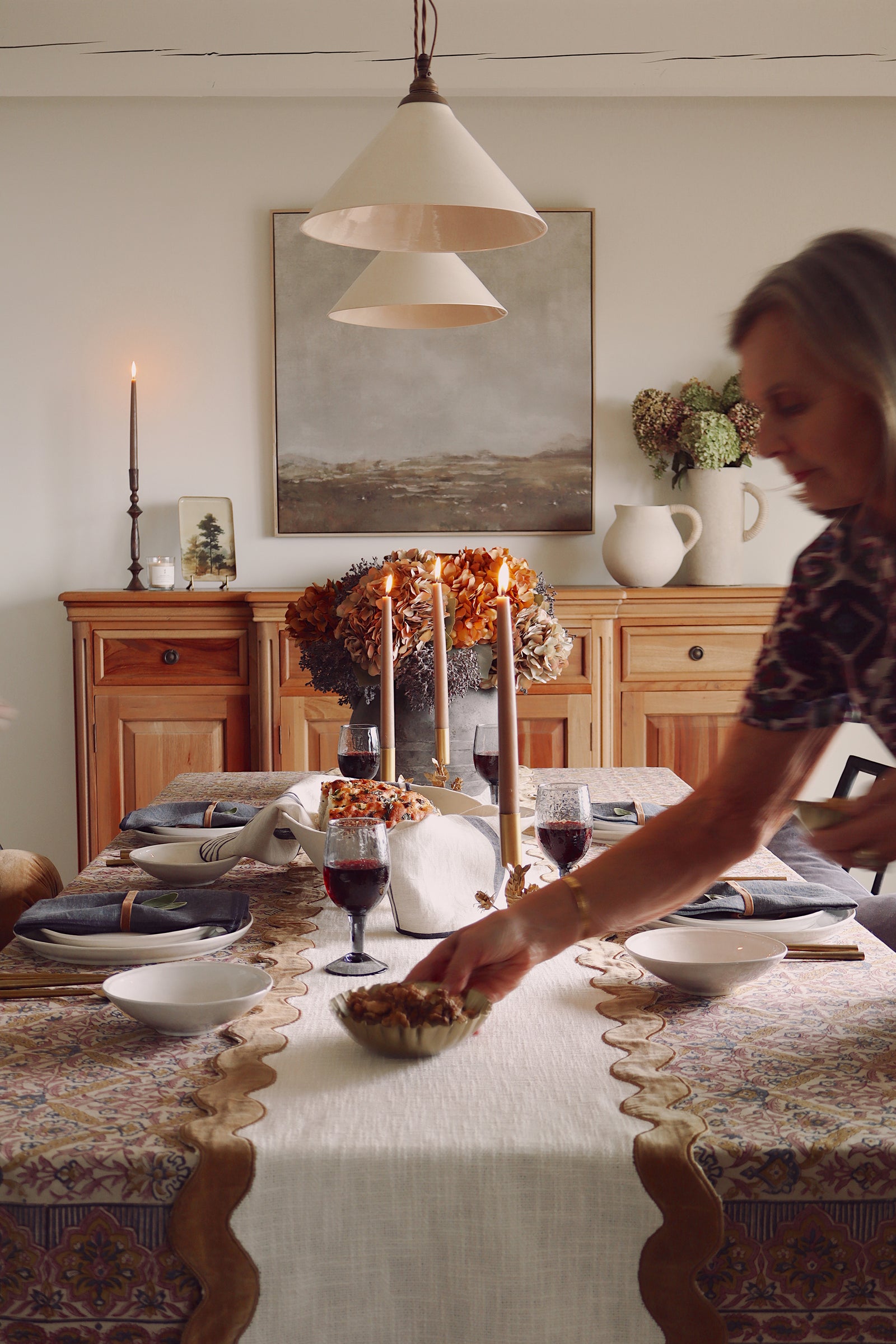 A woman in a patterned top is setting a table in a warm, rustic dining room. The table is decorated with a cream table runner, patterned tablecloths, wine glasses, and tall taper candles, with a wooden sideboard and a large landscape painting in the background.