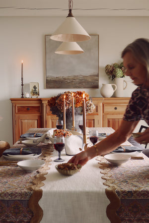 A woman in a patterned top is setting a table in a warm, rustic dining room. The table is decorated with a cream table runner, patterned tablecloths, wine glasses, and tall taper candles, with a wooden sideboard and a large landscape painting in the background.
