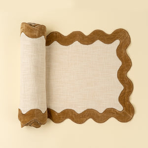 Rolled-up ivory table runner with gold scalloped velvet edge on a beige background. 