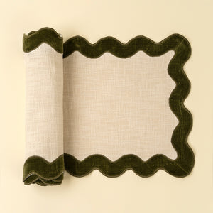 Rolled up table runner with scalloped forest green border, shown partially rolled on a light cream background. The cotton runner has a textured off-white linen center and a dark olive green velvet border.