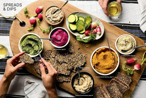 Assorted spreads and dips on a wooden board with bread and vegetables, hands holding a glass of white wine. "Scandinavian Everyday."