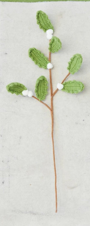 A crocheted green leafy branch with small white berries on a brown stem, laid flat on a light textured fabric background.