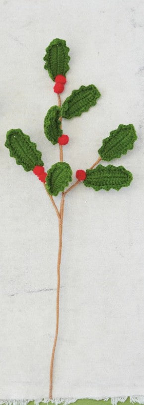 A crocheted holly branch decoration with green textured leaves, small red berries, and a long brown stem on a white fabric background.